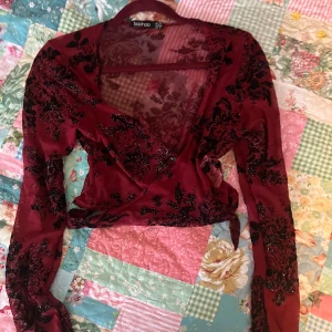 Vinröd glittrig omlottopp från Boohoo - Säljer en vinröd omlottopp från Boohoo med svarta glittriga blommönster. Köpt second hand men aldrig använd! Det står storlek L men skulle säga att den är i S/M