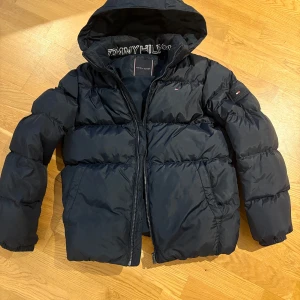 Svart dunjacka från Tommy Hilfiger - Säljer en svart dunjacka från Tommy Hilfiger med huva och dragkedja. Jackan har broderad logga på ärmen och stor logotyp på insidan av kragen. Perfekt för kyliga dagar.
