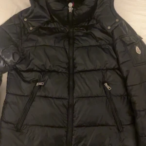 Svart pufferjacka från Moncler - Säljer en svart pufferjacka från Moncler med huva och dragkedja framtill. Jackan har två snedställda fickor med dragkedja och en ärmficka med Moncler-logga. Perfekt för kyliga dagar. Sommaren närmar sig och vill bli av med den så snabbt som möjligt 