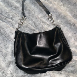 Svart axelväska med kedjedetaljer - Säljer en svart axelväska i glansigt material med silverfärgade kedjedetaljer på axelremmen. Väskan har dragkedja upptill och ett rymligt huvudfack. Perfekt för att bära dina viktigaste saker.