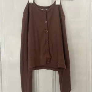 Brun ribbad kofta från 157 - Brun ribbad kofta från 157 i storlek M. Tröjan har långa ärmar, knappar framtill och är croppad i modellen. Perfekt att styla med jeans eller kjol för en avslappnad look.