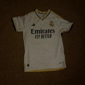 Real Madrid fotbollströja 2023/24 - Säljer en vit Real Madrid matchtröja från Adidas med svarta och gula detaljer. Tröjan har klubbmärke på bröstet och sponsortryck framtill. Klassisk V-ringning och korta ärmar. Perfekt för dig som älskar fotboll och Real Madrid.