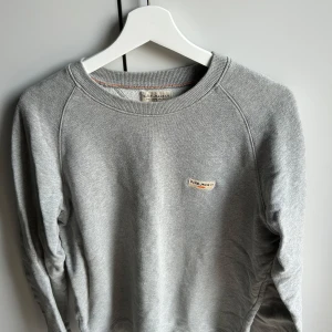 Grå sweatshirt från Nudie Jeans - Säljer en stilren grå sweatshirt från Nudie Jeans i 100% ekologisk bomull. Tröjan har rund halsringning, långa ärmar och en liten broderad logga på bröstet och i storlek M.