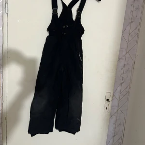 Svarta täckbyxor med hängslen från Tough 9 - Svarta täckbyxor från Tough 9 med justerbara hängslen och dragkedja framtill. Perfekta för kalla dagar och vinteraktiviteter. Broderad logga på benet och bred passform för extra rörelsefrihet.