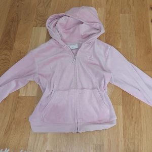Ljusrosa velour hoodie från Juicy Couture - Mysig ljusrosa hoodie i velour från Juicy Couture med dragkedja och huva. Jackan har broderad logga på bröstet och två fickor framtill. Perfekt för en avslappnad och trendig look.