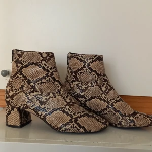 Ormskinnsmönstrade boots från Mango - Säljer ett par boots från Mango med ormskinnsmönster i beige och bruna toner. Skorna har en fyrkantig klack, rund tå och dragkedja bak. Perfekta för dig som vill sticka ut med din stil!