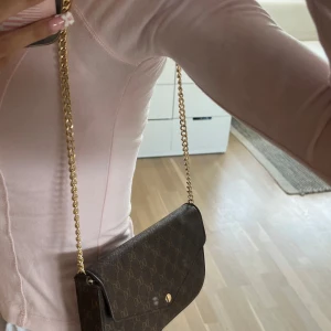 Brun axelväska med guldkedja från Don Donna - Säljer en brun axelväska från Glitter (Don Donna) med guldfärgad kedja och tryckknapp. Väskan har ett diskret mönster med DD-logga över hela ytan och ett innerfack med dragkedja. Perfekt för att bära det viktigaste med stil.