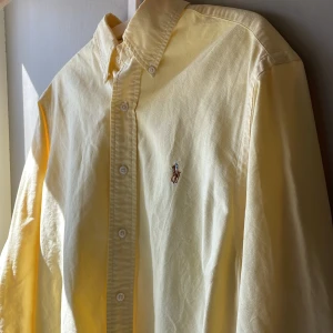 Gul skjorta från Ralph Lauren - Klassisk gul skjorta från Ralph Lauren med broderad logga på bröstet. Skjortan är tillverkad i 100% bomull och har knappar framtill samt lång ärm. Perfekt för en stilren och avslappnad look. Finns ett fåtal väldigt små ”fläckar” men som är så små att de inte kunde fångas på bild. Är osäker på exakt storlek men den är stor. Skriv till mig bara så skickar jag de mått du vill ha!😄