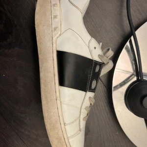 Valentino Garavani vita sneakers med svart detalj - Säljer ett par vita sneakers från Valentino Garavani med bred svart läderdetalj på sidan och vita snören. Skorna har rund tå och platt sula. Kommer med originalkartong och dustbag. Kan även gå med på att sänka priset här änvänt 1 månad bara lite smuts som går att ta bort med vatten skick8/10