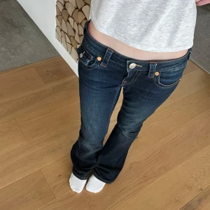 Mörkblå bootcut jeansbyxor från True Religion (Joey) - Säljer ett par mörkblå jeansbyxor från True Religion med bootcut passform och låg midja. Byxorna har tydliga kontrastsömmar, stora fickor med lock och silverfärgade knappar. Färgen har försvunnit lite längst ner på benet.