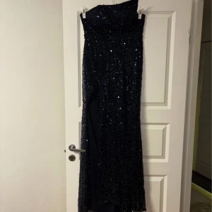 Marinblå glittrig långklänning från Cinderella Divine - Marinblå långklänning från Cinderella Divine med glittriga paljetter över hela klänningen. Klänningen har snörning i ryggen, är axelbandslös och har en slits framtill. Perfekt för bal eller festkvällar