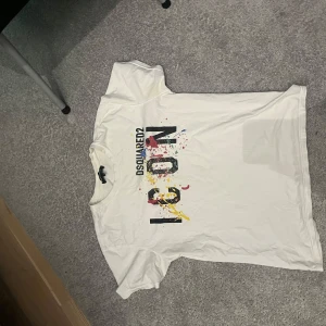 Vit t-shirt från Dsquared2 med färgglatt tryck - Säljer en vit t-shirt från Dsquared2 med stort färgglatt tryck och texten 'ICON' på bröstet. T-shirten har rund hals och korta ärmar. Perfekt för dig som gillar streetwear och vill sticka ut med en snygg design. Tröjan är i L men den sitter som M.