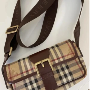 Rutig axelväska från Burberry - Säljer en klassisk axelväska från Burberry med det ikoniska rutiga mönstret i beige, svart, rött och vitt. Väskan har bruna skinn-detaljer, justerbar axelrem med Burberry-logga och ett snyggt spänne framtill. Perfekt för dig som gillar tidlös design.