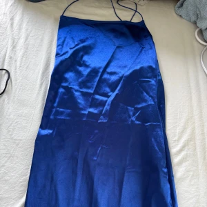 Blå satinklänning med smala axelband - Säljer en snygg blå satinklänning med smala axelband och rak halsringning. Klänningen har en slät, glansig yta och är figurnära upptill. Perfekt för dig som vill sticka ut med en elegant look.