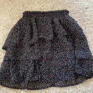 Svart volangkjol med mönster från H&M - Säljer en svart kjol från H&M med volanger och resår i midjan. Kjolen har ett småmönstrat tryck i vitt, rött och grönt och är tillverkad i ett lätt och luftigt material. Perfekt för dig som gillar en lekfull och trendig stil.