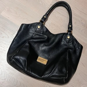 Svart handväska från Marc by Marc Jacobs - Snygg svart handväska i skinn från Marc by Marc Jacobs med guldfärgade metalldetaljer och dubbla handtag. Klassisk och rymlig modell med en liten metallbricka framtill. Perfekt för dig som vill ha en stilren accessoar.