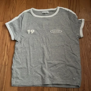 Grå t-shirt med vit text från Pull&Bear - Grå t-shirt från Pull&Bear med vita detaljer och tryck. Texten 'BE LIMITLESS NOW 1991' på ryggen och '19' framtill. Vit kant vid hals och ärmar. Perfekt för en avslappnad stil.