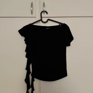 Svart topp med volangdetalj - Säljer en svart topp med kort ärm och volangdetalj längs ena sidan. Toppen har en enkel och stilren design som passar till många olika tillfällen.