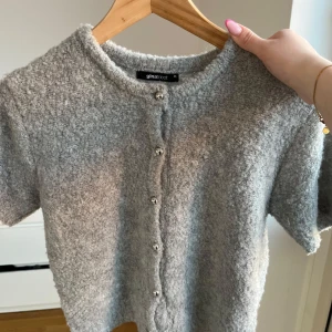 Grå kofta från Gina Tricot - Mysig grå kofta med korta ärmar från Gina Tricot. Tröjan har en fluffig, stickad känsla och knäpps framtill med pärlliknande knappar. Hör av dig vid fler frågor eller bilder❤️