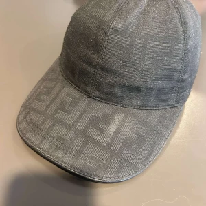 Svart keps från Fendi med diskret mönster - .Säljer en svart keps från Fendi med diskret mönster i tyget och justerbart spänne baktill i läder. Klassisk form med böjd skärm och broderad logga på spännet. Perfekt för dig som gillar stilrena accessoarer.pris kan diskuteras!!! Kontakta mig för mer info om kepsen.