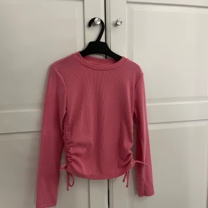 Rosa ribbad långärmad topp med snörning - Säljer en rosa långärmad topp med ribbad struktur och snörningsdetaljer i sidorna. Toppen har rund halsringning och är lätt croppad.