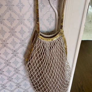 Beige nätväska med guldiga detaljer - Säljer en beige nätväska med guldiga kanter och handtag. Perfekt som accessoar till sommaren, lätt att bära och rymmer det viktigaste. Väskan har ett innerfoder i tyg och är både snygg och praktisk.