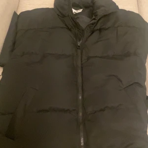 Svart dunväst från NA-KD - Säljer en svart dunväst från NA-KD med dragkedja framtill och hög krage. Västen har en enkel och stilren design, perfekt att bära över en hoodie eller tröja för extra värme. Hör av er vid funderingar eller frågor✅
