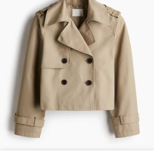 Beige croppad trenchcoat - Snygg croppad trenchcoat i beige med dubbelknäppning och klassisk krage. Jackan har långa ärmar med spännen vid ärmsluten och axelklaffar för en trendig look. Perfekt för vår och höst. Vill gärna byta mot en svart!!