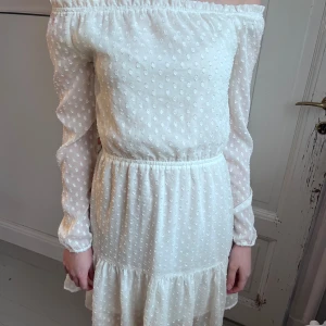 Vit offshoulder klänning med prickigt mönster - Superfin vit offshoulder klänning med långa ärmar och små prickar i tyget. Klänningen har resår i midjan och volang nertill för en söt look. Perfekt för sommarens tillfällen!