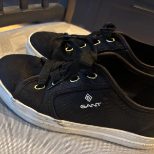 Svarta sneakers från GANT - Säljer ett par svarta sneakers från GANT med vita sulor och svarta snören. Skorna har guldfärgade öljetter och GANT-logga på sidan. Perfekta för en stilren och avslappnad look. Modellen på skorna heter Leisha Low Lace. Pris kan diskuteras 💕