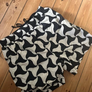 Svartvita mönstrade shorts från Bottega Veneta - Snygga shorts från Bottega Veneta med svartvitt grafiskt mönster. De har elastisk midja med snörning och är tillverkade i ett lätt syntetmaterial. Perfekta för sommardagar när du vill sticka ut lite extra.