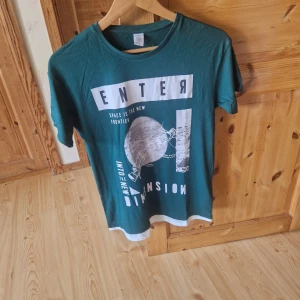 Grön t-shirt med tryck från Lindex - Säljer en grön t-shirt från Lindex med coolt vitt tryck framtill där det står 'ENTER' och motiv av en planet. T-shirten har rund hals och vita detaljer vid ärmslut och nederkant. Perfekt för dig som gillar rymdtema!