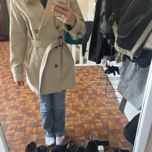 Beige trenchcoat från Aware - Snygg beige trenchcoat från Aware med klassisk dubbelknäppning och bälte i midjan. Jackan har långa ärmar och stilrena detaljer som knappar vid ärmslut och axlar. Perfekt för vår och höst.