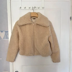 Beige teddyjacka med dragkedja - Mysig beige teddyjacka med stor krage och dragkedja framtill. Jackan är croppad och har lång ärm, perfekt för att hålla värmen under kyliga dagar. Passformen är normal och materialet är mjukt och fluffigt.