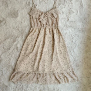 Romantisk sommarklänning från Guess - Söt blommig klänning, ljusbeige med små rosa blommor. Klänningen har smala axelband, volangdetaljer upptill och nertill samt pärlknappar framtill. 