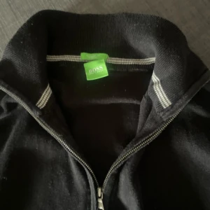 Svart zip upp från BOSS - Säljer en svart zip upp från BOSS med hög krage och diskret logga på bröstet. Den har dragkedja hela vägen och ribbade detaljer vid kragen. Perfekt för en stilren och avslappnad look. Mycket bra skick och en sällsynt gammal modellen green line av boss. Storlek M