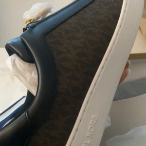 Svarta sneakers från Michael Kors - Säljer ett par snygga svarta sneakers från Michael Kors med diskret logomönster på sidan och vit platt sula. Skorna har en stilren design och är perfekta för dig som gillar klassiska märken.
