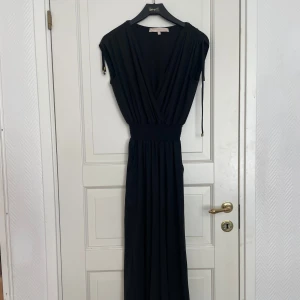 Svart jumpsuit med markerad midja - Säljer en svart jumpsuit med djup v-ringning och bred resår i midjan. Den har korta ärmar och ett elegant, flytande fall. Perfekt för dig som vill ha en stilren och tidlös look.