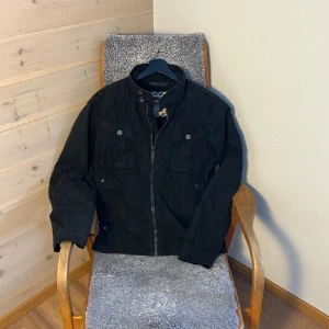 Svart jacka militärjacka  - Säljer en svart jacka med dragkedja framtill och ståkrage. Jackan har flera fickor med knappar och en stilren design. Perfekt för dig som vill ha en enkel och snygg jacka till vardags.