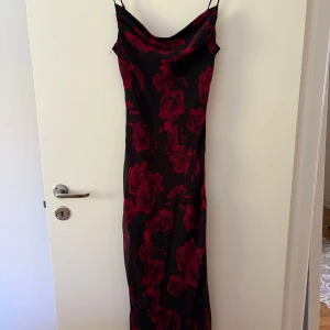 Svart långklänning med röda rosor från Gina Tricot - Säljer en svart långklänning från Gina Tricot med röda blommor. Klänningen har smala axelband och en snygg draperad urringning. Perfekt för dig som vill sticka ut med ett romantiskt mönster.