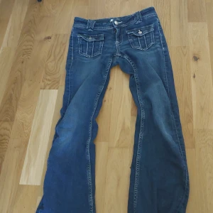 Blå bootcut jeans med fickdetaljer - Säljer ett par blå bootcut jeans med snygga fickor framtill som har lock och silverfärgade knappar. Jeansen har klassisk femficksdesign och markerade sömmar. Perfekta för dig som gillar en lite retro stil.