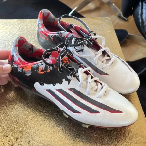 Adidas x Messi fotbollsskor  - Säljer ett par unika Adidas x Messi fotbollsskor i vitt med svarta och röda detaljer. Skorna har snörning och mönstrad yttersula för bra grepp på planen. Perfekta för dig som vill sticka ut på fotbollsplanen. Strl 40 (2/3)