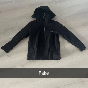 Svart skaljacka med huva från Arc'teryx - Svart skaljacka med huva från Arc'teryx. Jackan har dragkedja framtill, två sidofickor och justerbar huva. Perfekt för utomhusaktiviteter och skyddar mot vind och regn. Enkel och stilren design med logga på bröstet.