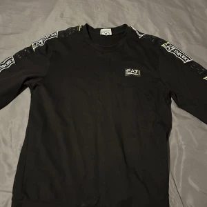 Svart långärmad tröja från EA7 Emporio Armani - Svart långärmad tröja från EA7 Emporio Armani med logga på bröstet och tryck längs ärmarna. Tröjan har en liten defekt med ett hål på ena ärmen. Perfekt för dig som gillar stilrena märkesplagg.