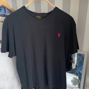 Svart v-ringad t-shirt från Polo Ralph Lauren - Svart t-shirt från Polo Ralph Lauren med v-ringning och rött broderat logotyp på bröstet. Klassisk passform och korta ärmar, perfekt för en stilren och enkel look.