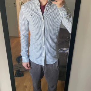 Vit och blå randig skjorta  - Klassisk vit och blå randig skjorta från Morris med knappar hela vägen och broderad logga på bröstet. Skjortan har lång ärm och normal passform, perfekt till många olika tillfällen. Den är i storlek M och i väldigt bra skick. Hör av er vid funderingar