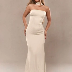 Champagnefärgad långklänning med halsdetalj - Säljer en elegant, figurnära långklänning i champagnefärg med unik halsdetalj och axelbandslös design framtill. Klänningen har en mjuk, slät yta och markerar midjan snyggt. Perfekt för speciella tillfällen.