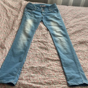 Blå jeansbyxor med knappar från Blue Ridge - Säljer ett par ljusblå jeansbyxor från Blue Ridge med tre knappar i midjan och klassiska fickor både fram och bak. Byxorna har en rak passform och snygga slitningar på låren för en trendig look.