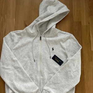 Ljusgrå hoodie från Polo Ralph Lauren - Säljer en ljusgrå hoodie från Polo Ralph Lauren med dragkedja och justerbar huva. Klassisk broderad logga på bröstet och två fickor framtill. Perfekt för en avslappnad stil.