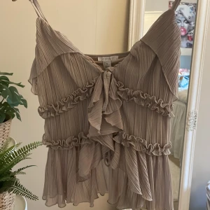 Beige plisserad linnetopp med volang från Amisu - Säljer en helt ny beige/grå volang topp från Amisu med volangdetaljer och knyt framtill. Tunt och luftigt material, perfekt för varma dagar. Smala axelband och romantisk känsla. Stl xl men skulle säga att den passar m-xl då den är ganska stretchig. Kan skicka fler bilder på om det önskas❤️
